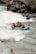 A group of four people is navigating through the rapids in a teal inflatable raft. They are all wearing safety gear, including helmets and life jackets. The river is turbulent with white water, and there are rocky cliffs and vegetation along the riverbank.