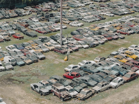 A large collection of old, abandoned cars parked closely together in an open field, many of which appear to be rusted or damaged. The vehicles are organized in loosely defined rows, and the ground is a mixture of grass and dirt. Some vegetation appears to be overgrowing parts of the cars.