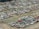 A large collection of old, abandoned cars parked closely together in an open field, many of which appear to be rusted or damaged. The vehicles are organized in loosely defined rows, and the ground is a mixture of grass and dirt. Some vegetation appears to be overgrowing parts of the cars.