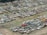 A large collection of old, abandoned cars parked closely together in an open field, many of which appear to be rusted or damaged. The vehicles are organized in loosely defined rows, and the ground is a mixture of grass and dirt. Some vegetation appears to be overgrowing parts of the cars.
