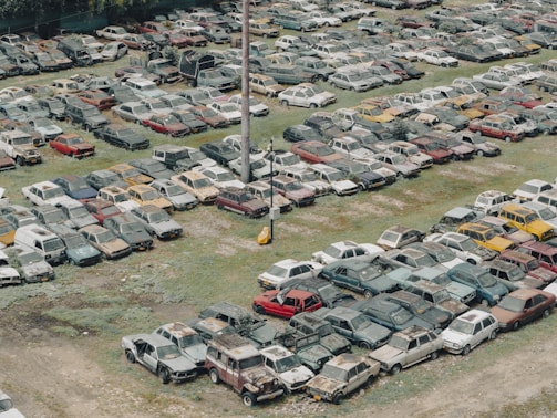 A large collection of old, abandoned cars parked closely together in an open field, many of which appear to be rusted or damaged. The vehicles are organized in loosely defined rows, and the ground is a mixture of grass and dirt. Some vegetation appears to be overgrowing parts of the cars.