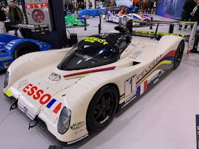 A sleek Peugeot race car is on display, featuring a white body adorned with colorful stripes and sponsorship logos such as Esso and Michelin. The car's aerodynamic design is highlighted by its low profile and large rear wing. Surrounding the car are other racing vehicles in a showroom environment with a crowd of people in the background.