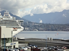 A large cruise ship is docked near a bustling cityscape with a backdrop of mountains under a cloudy sky. Numerous cars are parked near waterfront buildings, and a Canadian flag is visible, fluttering in the breeze.