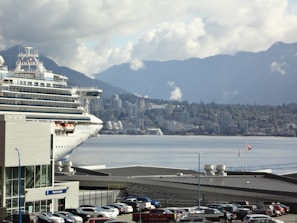 A large cruise ship is docked near a bustling cityscape with a backdrop of mountains under a cloudy sky. Numerous cars are parked near waterfront buildings, and a Canadian flag is visible, fluttering in the breeze.