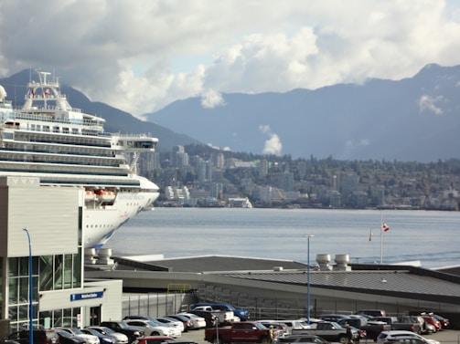 A large cruise ship is docked near a bustling cityscape with a backdrop of mountains under a cloudy sky. Numerous cars are parked near waterfront buildings, and a Canadian flag is visible, fluttering in the breeze.
