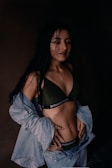 A woman with long dark hair is dressed in a dark green bralette with the word 'BRIGHTEST' on the band and light blue denim jeans and jacket. Her hands are on her hips, and she seems to have damp hair, suggesting she might have been in the rain or just out of the shower. The background is dark, creating a high contrast with her clothing and skin.