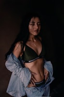 A woman with long dark hair is dressed in a dark green bralette with the word 'BRIGHTEST' on the band and light blue denim jeans and jacket. Her hands are on her hips, and she seems to have damp hair, suggesting she might have been in the rain or just out of the shower. The background is dark, creating a high contrast with her clothing and skin.