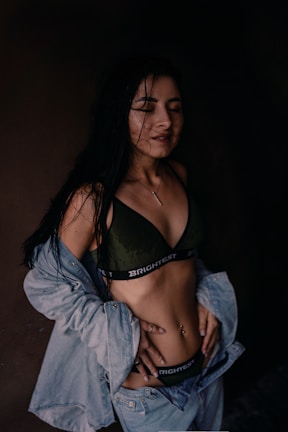 A woman with long dark hair is dressed in a dark green bralette with the word 'BRIGHTEST' on the band and light blue denim jeans and jacket. Her hands are on her hips, and she seems to have damp hair, suggesting she might have been in the rain or just out of the shower. The background is dark, creating a high contrast with her clothing and skin.
