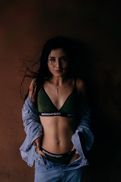 A woman stands against a dark brown background, wearing a dark green bra with 'BRIGHTEST' written on the band and an unbuttoned denim jacket. The lighting is low, creating dramatic shadows, and her long dark hair is loose, partially covering her face.
