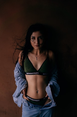 A woman stands against a dark brown background, wearing a dark green bra with 'BRIGHTEST' written on the band and an unbuttoned denim jacket. The lighting is low, creating dramatic shadows, and her long dark hair is loose, partially covering her face.