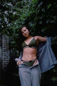 A woman stands in a lush outdoor setting, surrounded by dense greenery. She is dressed in a green bikini top and jeans, partially covered by an unbuttoned blue denim shirt. Her long dark hair falls over her shoulders, and she looks directly at the camera.