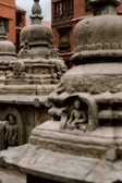 Ancient stone stupas adorned with intricate carvings stand in an outdoor setting. The focus is on the detailed architecture of the stupas, including carved figures in recessed niches. A background of brick buildings adds to the historical atmosphere.