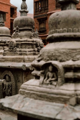 Ancient stone stupas adorned with intricate carvings stand in an outdoor setting. The focus is on the detailed architecture of the stupas, including carved figures in recessed niches. A background of brick buildings adds to the historical atmosphere.