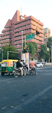 A city street with a group of motorcycles and an auto-rickshaw waiting at a traffic signal. The background features a large pink building with multiple stories and distinctive architecture. There are lush green trees and streetlights around the intersection.