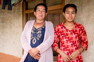 Two women are standing side by side in front of a rustic wall, smiling warmly. They are wearing traditional clothing with intricate patterns and vibrant colors. The woman on the left is dressed in a dark blue outfit with gold embroidery and a light pink cardigan, while the woman on the right is in a red dress with white geometric patterns. Both appear relaxed and content.
