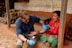 A healthcare professional wearing blue gloves and a dark blue shirt is checking the blood pressure of an elderly woman using a stethoscope and a blood pressure monitor. The woman is seated on a porch, dressed in traditional attire with a red blouse and green skirt. There are wooden pillars and a rustic background of a lightly white-washed wall with brown patches behind them.