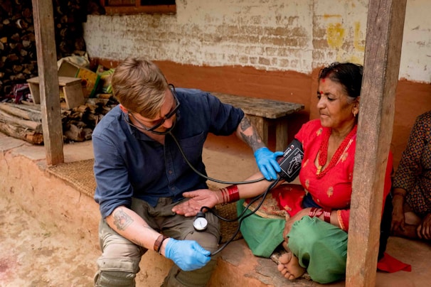 A healthcare professional wearing blue gloves and a dark blue shirt is checking the blood pressure of an elderly woman using a stethoscope and a blood pressure monitor. The woman is seated on a porch, dressed in traditional attire with a red blouse and green skirt. There are wooden pillars and a rustic background of a lightly white-washed wall with brown patches behind them.