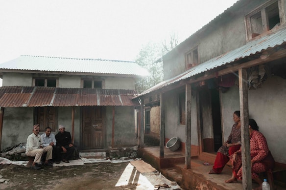 Two residential houses with sloping corrugated metal roofs are shown. In front of one house, three men are seated together while two women sit on the porch of the adjacent house. The houses have a rustic appearance with wooden doors and a few visible windows.