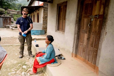 A man wearing sunglasses and a stethoscope stands outdoors, putting on blue gloves, while a woman sits on a porch step nearby, dressed in traditional clothing. The setting appears to be a rural area with old, simple buildings and rustic surroundings.