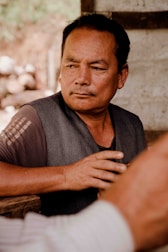 A middle-aged man with short dark hair and a contemplative expression, sitting at a wooden table. He is wearing a dark gray vest over a brown shirt, and his hand is resting on the table. The background is blurred, suggesting an outdoor setting with a rustic feel.