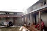 A rural scene depicting two small houses with corrugated metal roofs and several people sitting outside. One group of three individuals is sitting on the left near the mud-brick house, while two women sit on the right porch. The environment appears to be rustic with earthy tones dominating the setting and greenery partially visible in the background.