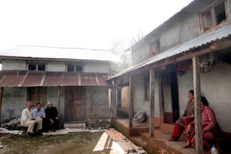 A rural scene depicting two small houses with corrugated metal roofs and several people sitting outside. One group of three individuals is sitting on the left near the mud-brick house, while two women sit on the right porch. The environment appears to be rustic with earthy tones dominating the setting and greenery partially visible in the background.