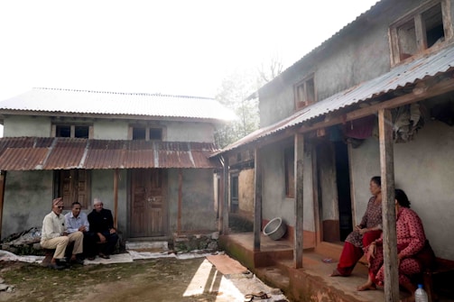 A rural scene depicting two small houses with corrugated metal roofs and several people sitting outside. One group of three individuals is sitting on the left near the mud-brick house, while two women sit on the right porch. The environment appears to be rustic with earthy tones dominating the setting and greenery partially visible in the background.