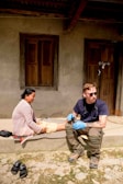 A person wearing sunglasses and medical gloves sits on a step next to a woman, who appears to be receiving medical attention. The setting is outdoors in front of a building with wooden doors and windows. There are sandals placed on the ground nearby.