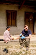 A person wearing sunglasses and medical gloves sits on a step next to a woman, who appears to be receiving medical attention. The setting is outdoors in front of a building with wooden doors and windows. There are sandals placed on the ground nearby.