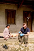A person wearing sunglasses and medical gloves sits on a step next to a woman, who appears to be receiving medical attention. The setting is outdoors in front of a building with wooden doors and windows. There are sandals placed on the ground nearby.