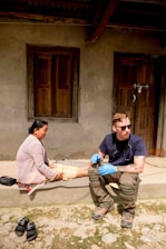 A person wearing sunglasses and medical gloves sits on a step next to a woman, who appears to be receiving medical attention. The setting is outdoors in front of a building with wooden doors and windows. There are sandals placed on the ground nearby.
