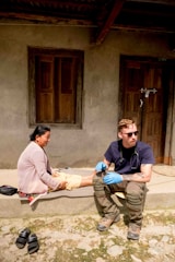 A person wearing sunglasses and medical gloves sits on a step next to a woman, who appears to be receiving medical attention. The setting is outdoors in front of a building with wooden doors and windows. There are sandals placed on the ground nearby.