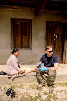 A person wearing sunglasses and medical gloves sits on a step next to a woman, who appears to be receiving medical attention. The setting is outdoors in front of a building with wooden doors and windows. There are sandals placed on the ground nearby.