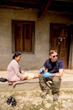 A person wearing sunglasses and medical gloves sits on a step next to a woman, who appears to be receiving medical attention. The setting is outdoors in front of a building with wooden doors and windows. There are sandals placed on the ground nearby.