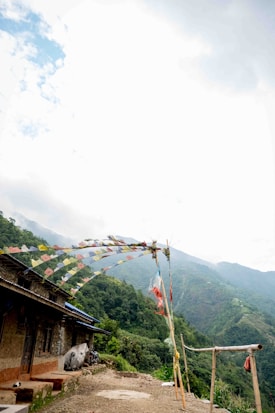 A rustic building sits on a hillside overlooking lush green mountains. Colorful prayer flags are strung across poles, fluttering in the wind. The sky is partially cloudy.