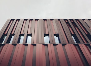 A modern building facade featuring vertical metal panels in varying shades of red and brown. The design is geometric and repetitive, with windows interspersed between the panels. The sky above is cloudy and gray, contributing to a muted overall appearance.