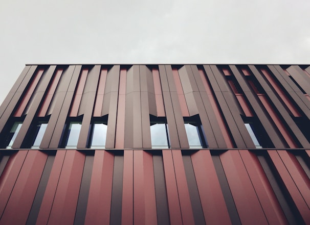 A modern building facade featuring vertical metal panels in varying shades of red and brown. The design is geometric and repetitive, with windows interspersed between the panels. The sky above is cloudy and gray, contributing to a muted overall appearance.