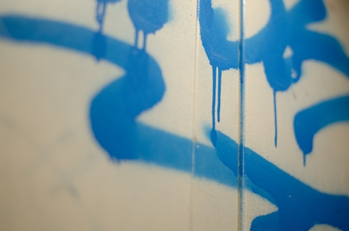 Blue spray paint drips on a light-colored wall create an abstract pattern with soft shadows.