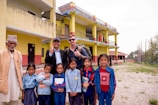 A group of people, including children and adults, stand outdoors in front of a yellow and red building. The children are in school uniforms, holding books, and some adults are smiling and making peace signs. The setting appears to be a school, with flags visible on the building and an open area with grass in the foreground.