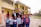 A group of people, including children and adults, stand outdoors in front of a yellow and red building. The children are in school uniforms, holding books, and some adults are smiling and making peace signs. The setting appears to be a school, with flags visible on the building and an open area with grass in the foreground.