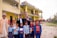 A group of people, including children and adults, stand outdoors in front of a yellow and red building. The children are in school uniforms, holding books, and some adults are smiling and making peace signs. The setting appears to be a school, with flags visible on the building and an open area with grass in the foreground.