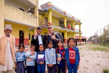 A group of people, including children and adults, stand outdoors in front of a yellow and red building. The children are in school uniforms, holding books, and some adults are smiling and making peace signs. The setting appears to be a school, with flags visible on the building and an open area with grass in the foreground.