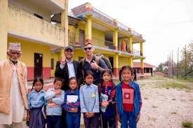 A group of people, including children and adults, stand outdoors in front of a yellow and red building. The children are in school uniforms, holding books, and some adults are smiling and making peace signs. The setting appears to be a school, with flags visible on the building and an open area with grass in the foreground.