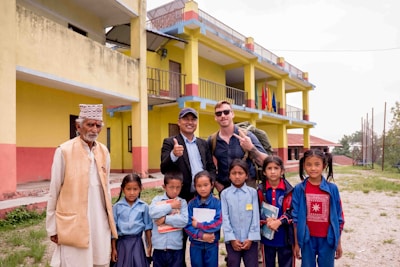 A group of people stands in front of a colorful building with two stories and several balconies. The group includes an elderly man in traditional attire, two men with backpacks giving a thumbs up or peace sign, and several children in school uniforms holding notebooks. The setting appears to be a school or educational facility in a rural area, with grass and trees in the background.