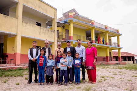 A group of adults and children are standing in front of a colorful two-story school building with balconies and flags. The adults are dressed in formal and traditional attire, and the children are wearing school uniforms.