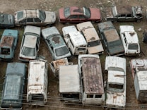 A collection of old, abandoned cars and vans are tightly packed in an outdoor area. The vehicles are covered in rust and dirt, and many have missing windows or peeling paint. Overgrown vegetation peeks through some of the gaps, adding to the sense of decay.