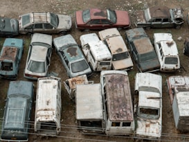 A collection of old, abandoned cars and vans are tightly packed in an outdoor area. The vehicles are covered in rust and dirt, and many have missing windows or peeling paint. Overgrown vegetation peeks through some of the gaps, adding to the sense of decay.