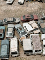 An assortment of old, rusted cars and vans is parked haphazardly on a patch of dirt and gravel. The vehicles show signs of decay with peeling paint and visible rust. Some cars are stacked closely, and their windows are either broken or missing. The surrounding area is barren, with scattered small rocks and patches of grass.