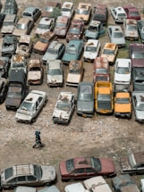 An aerial view of an outdoor area filled with numerous old and rusted cars parked closely together. The cars display a range of colors and visible signs of decay and neglect, such as peeling paint and rust. A person wearing dark clothing and a hat is walking on the rocky ground near the bottom of the image.