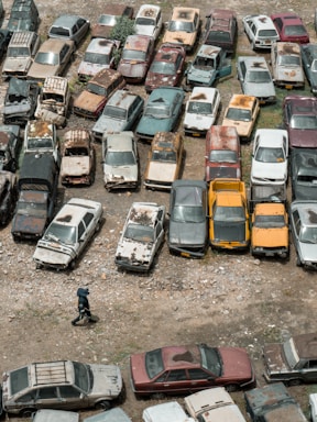 An aerial view of an outdoor area filled with numerous old and rusted cars parked closely together. The cars display a range of colors and visible signs of decay and neglect, such as peeling paint and rust. A person wearing dark clothing and a hat is walking on the rocky ground near the bottom of the image.